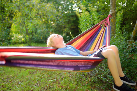 Cute preteen blond boy having fun with multicolored hammock in backyard or outdoor playground. Summer outdoors active leisure for kids. Child relaxing and swinging in hammock.の写真素材