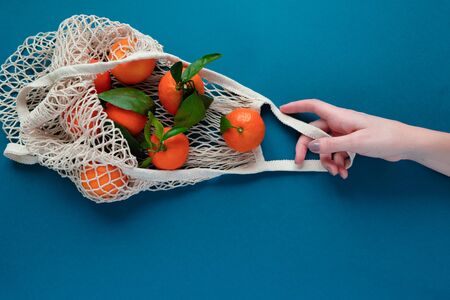 A human hand holds a string bag with tangerines on blue classic color background. Eco concept, healthy food, vitamins.の写真素材