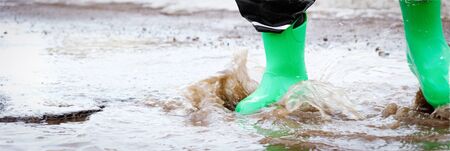 child with green rain boots jumps into a puddle. Long format for webの写真素材