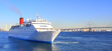 white cruise ship sailing with blue sky, sea water. bridge and cityscape in the back. 2.21:1 ratio, room for text. yokohama port tokyo bay japanの写真素材