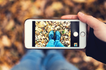 The girl makes a photo sneakers on a background of colorful dry autumn leaves, on her smartphoneの写真素材