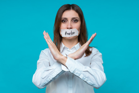 Closeup portrait of young caucasian brown-haired girl demonstrates by hands the prohibition sign, the mouth is sealed with a white plaster with an inscription #metoo. Text space. #metoo movement concept.の写真素材