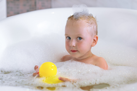 Adorable bath baby boy with soap suds on hair play with colorful toy yellow duck. Hygiene and care for young children.の写真素材