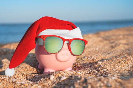 Toy piggy bank in form of pink pig in sunglasses with polarizing lenses and Christmas Santa hat is standing on sandy beach on sunny day, symbols of years of the eastern calendar, Chinese zodiacの写真素材