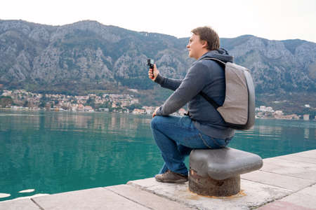 A handsome young guy, a travel blogger, takes his trip to an action camera, sits on the seashore with beautiful views of the mountains.の写真素材