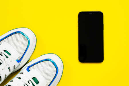 Flat top on white sneakers with blue and green decorations and black mobile phone lying on bright yellow background. Walking or hiking concept, copy space for any text or advertisement.の写真素材