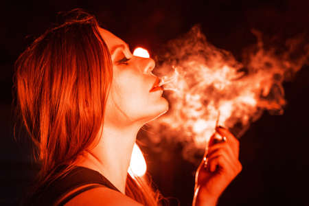 Young caucasian woman smoking in back light witn electric red color tone. ? lose up portrait on dark background.の写真素材