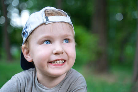 Cute little caucasian baby boy with blond hair and blue eyes smiling. Outdoor, green park or forest around there. Casual wear, cap backwards.の写真素材