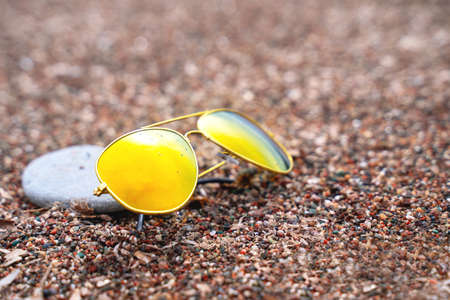 Sunglasses with polarizing lenses with pebble nearby lie on sand, close up. Protection against harmful ultraviolet radiation from sun. Accessories for vacation trip to warm countries to sea or ocean.の写真素材