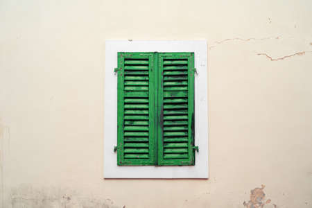 Old closed green wooden shutters on light plastered wall with cracks and time spots, in...
