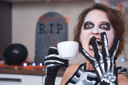 Young contented woman with creepy makeup and in skeleton costume drinks refreshing, relaxing or hot beverage from mug at Halloween party, decorated apartment.の写真素材