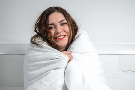 Attractive young brunette woman has wrapped herself in white duvet to keep warm and is laughing, while sitting on the bed. She feels the approach of winter. Lack of heating at home in cold season.の写真素材