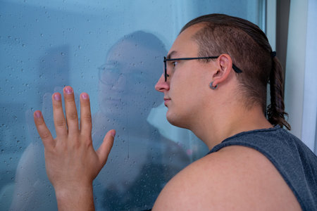 Young man with ponytail and in glasses looks sadly out of window, putting his hand on the glass. He...