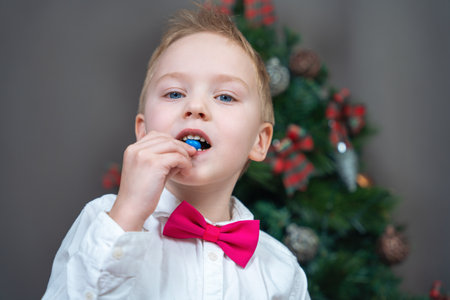 Lovely boy in festive outfit with shirt and pink bow tie eats candy in blue glaze. Decorated...
