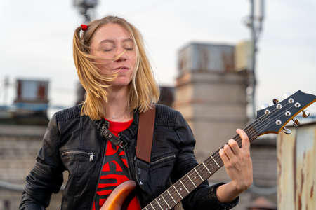 Young rock blonde girl with two funny ponytails, closed eyes , playing an electric guitar on the roof of an old building, front view. Musical video clip shooting. Outdoor show.の写真素材
