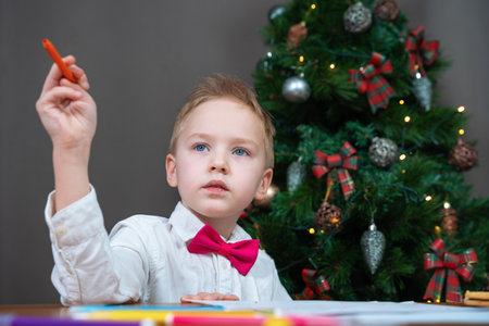 Child in festive outfit with bow tie sits thoughtfully with pen in his hand and writes letter to Santa about gifts, or does school homework during the Christmas holidays.の写真素材