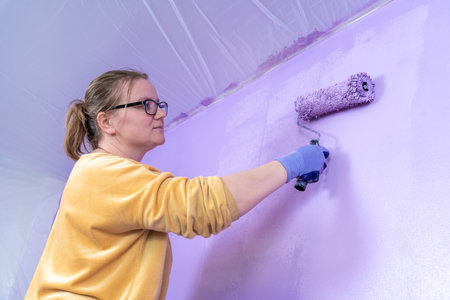 Woman in glasses paints wall in lilac color, holds roller with her hand in glove. Self-designer decorates wall, makes repairs, renews the shade.の写真素材