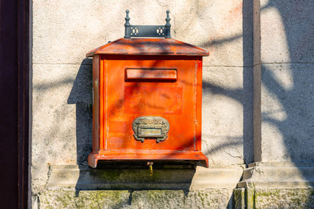 An ornate vintage red mailbox adorns a textured stone wall. shadows play across the surface,...