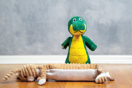 A charming scene featuring a green plush dinosaur dog toy standing next to a lying floppy stuffed bunny on a wooden floor with a textured gray wall in the background.の写真素材