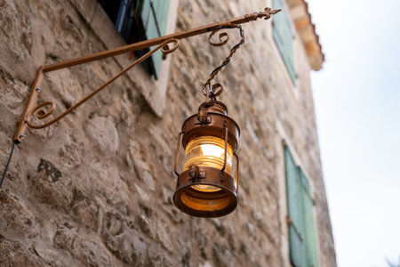 A classic lantern illuminates a rustic stone wall, featuring green shutters, evoking a nostalgic and charming atmosphere in an old town setting.の写真素材