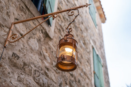 A warm, glowing lantern hangs on an old stone wall beside green shutters, reflecting rustic charm and classic architecture. perfect for themes of history, light, and vintage design.の写真素材