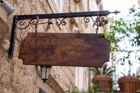 A rustic wooden sign hangs empty from an ornate iron bracket in a charming vintage street. the backdrop features stone walls and potted plants, creating a quaint atmosphere.の写真素材