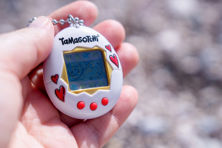 Bar, Montenegro: September 24, 2025: A close-up of a persons hand holding a white Tamagotchi with red hearts and buttons, featuring a blue screen and a chain, against a blurred outdoor backgroundのeditorial素材
