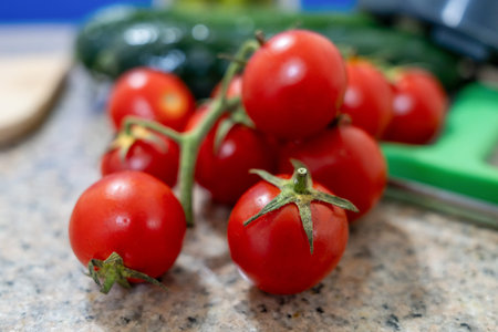 A fresh cluster of red cherry tomatoes still on the vine sits on a speckled kitchen counter next to green cucumbers and a green cutting board in a bright kitchenの写真素材