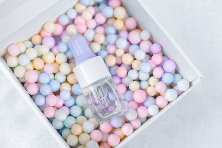 Clear glass dropper bottle with lavender cap lying in a white box filled with pastel foam balls, minimal skincare or cosmetic product flat lay with soft colors and clean background copy spaceの写真素材