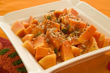 Freshly cut papaya cubes with mixed sunflower, pumpkin and sesame seeds on a square white plate with on an orange tableclothの写真素材