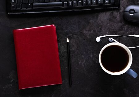 Office desk with notebook, mug of coffee or tea, keyboard, mouse, headphones and pencil on blackの写真素材