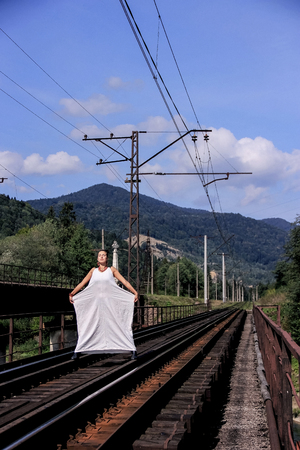 girl in a white dress on a blue sky background. Young woman on the background of the green mountains of Georgia. The lady is standing on the iron rails. Waiting for the train. Journey through Georgia.の写真素材