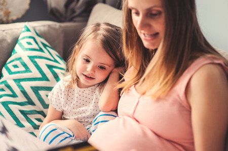 happy pregnant mother and toddler daughter playing and reading book at home on couch, cozy lifestyle captureの写真素材