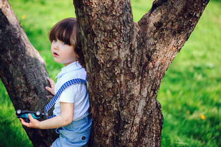 cute toddler child boy with long hair in stylish outfit playing with toy car on the walk in summer or sping garden, sitting on big treeの写真素材