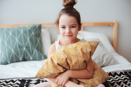 happy child girl sitting on bed and hugs pillow, waking up in early morning or going to sleep. Kid in comfortable room enjoying weekendの写真素材