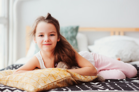 happy little child girl lying on her bed in the morning, waking up in comfortable room with modern bedlinen and pillows, cozy homely scene の写真素材