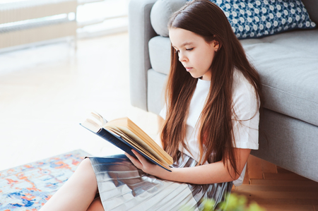 smart kid girl reading interesting book  at home, sitting with couch on background. Learning and education conceptの写真素材