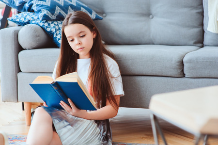 smart kid girl reading interesting book  at home, sitting with couch on background. Learning and education conceptの写真素材