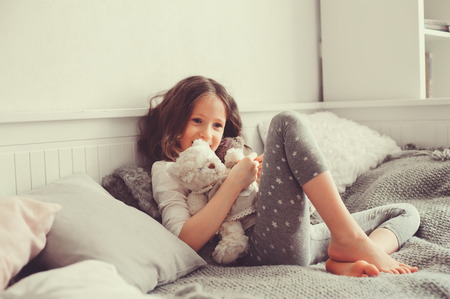 happy kid girl playing with teddy bears in her room, sitting on bed in pajamaの写真素材