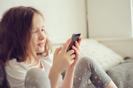 kid girl playing smartphone at home on the bedの写真素材