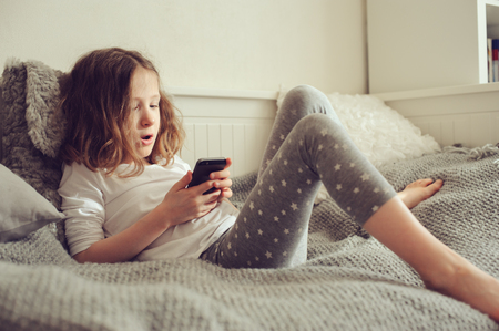 kid girl playing smartphone at home on the bedの写真素材