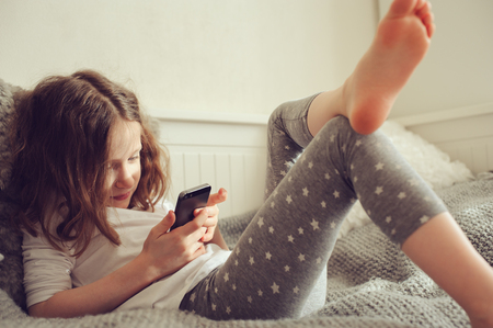 kid girl playing smartphone at home on the bedの写真素材