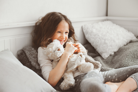 happy kid girl playing with teddy bears in her room, sitting on bed in pajamaの写真素材