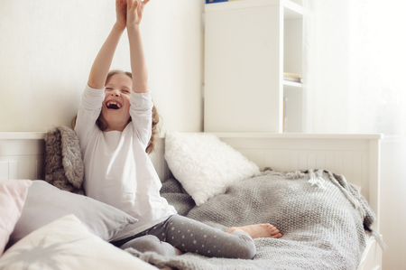 happy kid girl stretching in her room, sitting on bed in the morningの写真素材