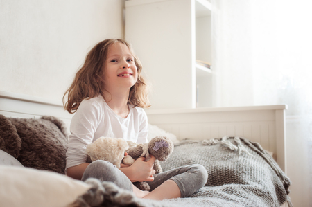 happy kid girl playing with teddy bears in her room, sitting on bed in the morningの写真素材