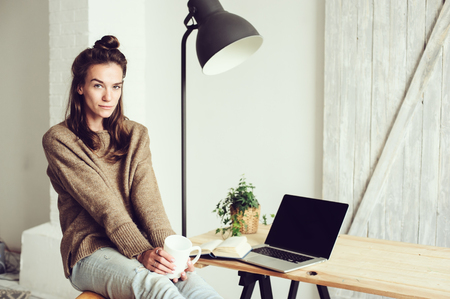 young beautiful woman shopping at home online with laptop and cup of coffee in the morning. Home office in modern scandinavian interiorの写真素材