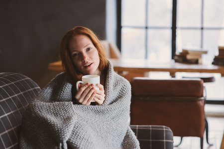 young sick woman healing with hot drink at home on cozy couch, wrapped in knitted blanketの写真素材