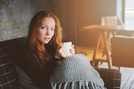 young sick woman healing with hot drink at home on cozy couch, wrapped in knitted blanketの写真素材