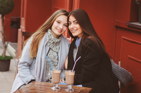two happy girl friends talking and drinking coffee in autumn city in cafe. Meeting of good friends, young fashionable students with natural make up.の写真素材