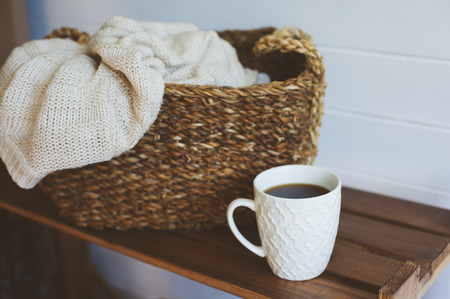 cozy winter interior details in white and brown tones. Basket with knitted sweater and cup of coffee on wooden shelf. の写真素材